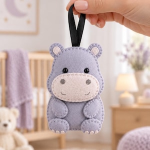 May include: A handcrafted felt hippo ornament in shades of lavender and light pink. The hippo features black bead eyes, a stitched mouth, and small rounded ears. A black ribbon loop is attached to the top for hanging, with visible blanket stitching.
