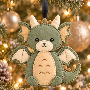 May include: A felt dragon ornament in shades of sage green and cream, featuring stitched details, two small horns, wings, and a curved tail. The creature has black bead eyes and a stitched nose and mouth, suspended by a black ribbon loop.