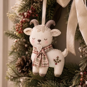 May include: A felt goat ornament in white with grey horns and hooves, featuring closed eyes, pink cheeks, and a patterned scarf with red and green floral details. The goat has a small embroidered sprig on its side and hangs from a cream ribbon.