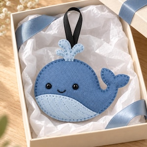 Op de afbeelding: Een blauwe walvisornament van vilt met een lichtblauwe buik, een kleine waterstraal bovenop en twee zwarte kraaloogjes. De walvis heeft een gestikte omtrek en een zwart lintje om op te hangen, gepresenteerd in een crèmekleurige geschenkdoos met vloeipapier.