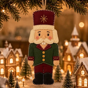 May include: A felt nutcracker ornament featuring a maroon hat with a gold star, a green jacket with three yellow buttons, maroon trousers, and black boots. The figure has a white mustache, visible teeth, and pink cheeks, suspended by a twine loop.