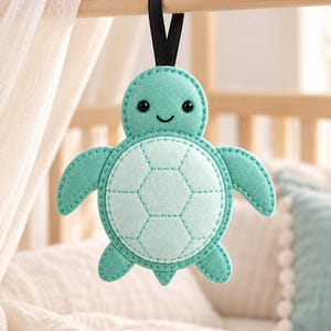 Könnte beinhalten: Ein Filz-Anhänger in Form einer Meeresschildkröte in Mint- und Hellgrüntönen. Die Schildkröte hat ein aufgesticktes hexagonales Panzer-Muster, zwei kleine schwarze Perlenaugen und ein aufgesticktes Lächeln. Eine schwarze Schlaufe dient zum Aufhängen.