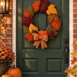 May include: A circular wreath constructed from felt leaves in shades of orange, yellow, olive green, and deep burgundy. The wreath features a central orange felt pumpkin, a brown felt acorn, and a large tan felt bow at the bottom base.
