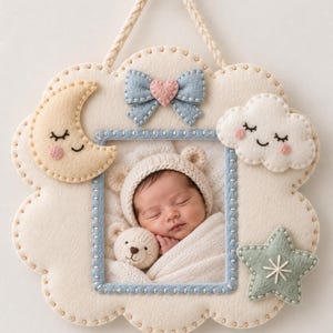 Puede incluir: Un marco de fotos de fieltro color crema con borde festoneado y cordón trenzado para colgar. El marco incluye una foto de un bebé durmiendo, una luna creciente amarilla, un lazo azul con un corazón rosa, una nube blanca y una estrella verde salvia.