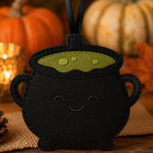 May include: A black felt cauldron ornament featuring a green felt liquid top with circular bubbles and a stitched smiling face. The cauldron has two side handles, three small feet, and a black ribbon loop attached to the top for hanging purposes.