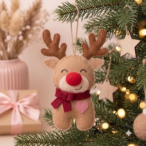 May include: A felt reindeer ornament featuring a tan body, dark brown antlers, a bright red nose, and a dark red scarf. The reindeer has a small pink heart stitched onto its chest and is suspended by a thin twine loop for hanging on a tree.