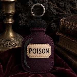 May include: A dark purple felt keychain shaped like a bottle with a black felt stopper and a silver metal ring. A beige rectangular label is stitched onto the center with the word POISON embroidered in black thread, and a black crescent shape.
