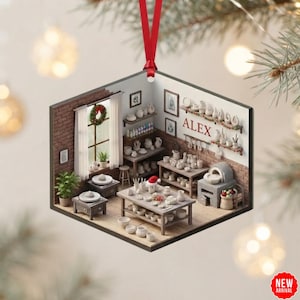 May include: A square-shaped hanging ornament featuring a miniature pottery studio scene with brick walls, wooden shelves, a kiln, and various ceramic pieces. The name ALEX is printed in red on the wall above a shelf. A red ribbon is attached to the top.
