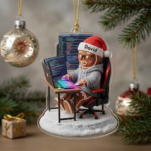 May include: A clear acrylic Christmas ornament featuring a gingerbread person wearing a grey hoodie, glasses, and a red Santa hat with the name David printed on it. The figure sits in a black and red gaming chair at a desk with a laptop.
