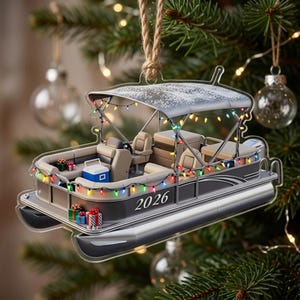 May include: A flat, transparent acrylic Christmas tree ornament shaped like a pontoon boat. The boat features tan seating, a blue cooler, and colorful string lights. The side of the boat is printed with the year 2026 and small gift boxes.