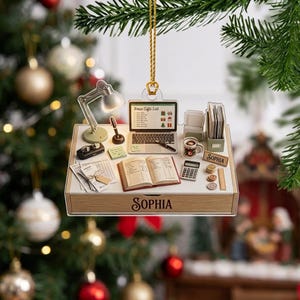 May include: A flat, rectangular hanging ornament featuring a detailed desk scene with a laptop displaying 'Xmas Gifts List', an open book, a lamp, a mug, a calculator, and coins. The front edge of the desk is wood-toned and printed with the name 'SOPHIA'.