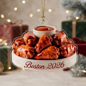 May include: A clear, flat, bell-shaped ornament featuring a pile of glossy, red-sauced chicken wings on a white circular base. A small white bowl of red dipping sauce sits atop the wings. The text Boston 2026 is printed in red on the base.