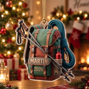 May include: A clear acrylic hanging ornament shaped like a hiking backpack with green and brown panels, brown straps, and black buckles. It features two crossed trekking poles, a coiled blue climbing rope, and a red water bottle. Text reads MARTIN 2026.