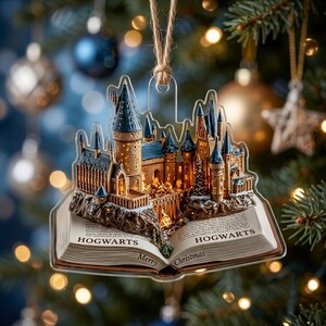 May include: A clear acrylic Christmas ornament shaped like an open book with a three-dimensional Hogwarts castle model emerging from the pages. The text HOGWARTS and Merry Christmas is printed on the open pages, suspended by a brown twine string.