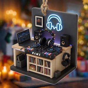 May include: A hanging ornament featuring a miniature DJ studio setup with a laptop, a mixing console labeled NAME, two speakers, and two pairs of headphones. The dark backdrop displays a glowing neon-style headphone icon and a small framed picture.