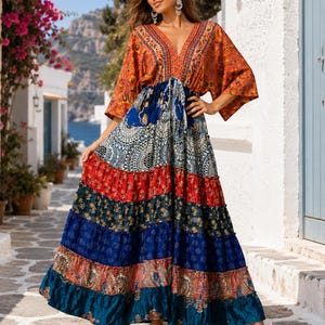 May include: A floor-length maxi dress featuring a deep V-neckline and wide kimono-style sleeves. The bodice displays an orange paisley print, while the skirt consists of tiered horizontal panels in contrasting patterns of blue, red, and teal.