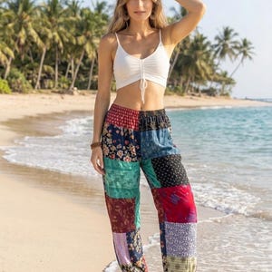Może przedstawiać: Biały bezrękawnik typu crop top ze sznurkiem ściągającym pośrodku, zestawiony z luźnymi spodniami typu patchwork. Spodnie składają się z różnorodnych kolorowych paneli materiałowych z motywami kwiatowymi, geometrycznymi i paisley w odcieniach czerwieni, błękitu i zieleni.