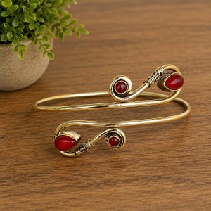 May include: A gold-toned metal arm cuff featuring a bypass design with two open ends. Each end terminates in a spiral detail accented with a smooth, deep red, teardrop-shaped stone and a smaller, circular red stone set within the metal wire.