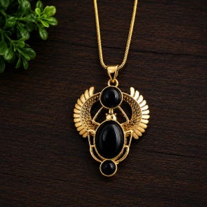May include: A gold-toned necklace featuring a pendant shaped like a winged scarab beetle. The pendant is accented with three smooth, oval-shaped black stones set into the metal frame. The piece hangs from a matching gold-toned snake chain.