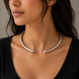 Könnte beinhalten: Ein silberfarbenes Metall-Halsband in Form eines glatten, starren und offenen Rings. Die Kette endet vorne mit zwei kleinen, polierten kugelförmigen Akzenten, was für ein schlichtes und klares Erscheinungsbild sorgt.