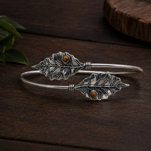 Può includere: Un bracciale aperto color argento con due estremità dettagliate a forma di foglia. Ogni foglia presenta una superficie strutturata con venature prominenti ed è impreziosita da una piccola pietra preziosa rotonda occhio di tigre marrone.