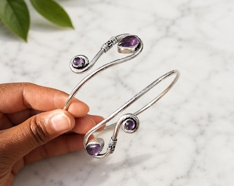 May include: A silver-toned bypass cuff bracelet featuring two open ends that spiral into decorative silver coils. Each end is accented with a small, round purple amethyst stone and a larger, pear-shaped purple amethyst stone set in silver.