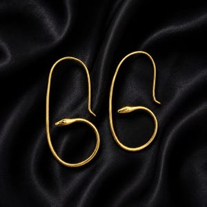 May include: A pair of gold-toned earrings shaped like elongated loops with a snake head motif at the base of each loop. The snake heads feature small, defined eyes and a smooth, curved body that forms the bottom section of the earring structure.