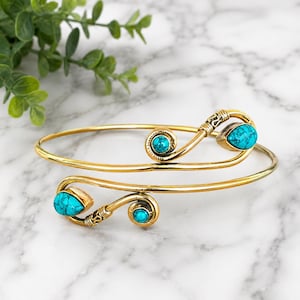 May include: A gold-toned metal arm cuff bracelet featuring a bypass design with two spiraled ends. Each end is accented with a teardrop-shaped turquoise stone and a smaller round turquoise stone, all set in smooth gold-toned metal bezels.