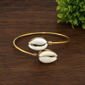 Puede incluir: Una pulsera rígida de metal en tono dorado con dos conchas de cauri blancas en los extremos abiertos. Las conchas están aseguradas a la fina banda de alambre pulido de color dorado con pequeñas envolturas de alambre, creando un diseño abierto.