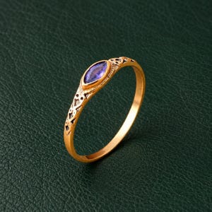 Op de afbeelding: Een gouden ring met een paarse edelsteen in marquise-slijpvorm in het midden. De ringband is voorzien van een ingewikkeld, gestructureerd patroon langs de schacht, dat zich uitstrekt van de centrale zetting tot aan de zijkanten van de dunne ring.