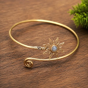 May include: A gold-toned metal cuff bracelet featuring an open design with a sun-shaped ornament at one end. The center of the sun motif contains a small, round, iridescent blue stone. The opposite end of the bracelet terminates in a spiral coil.