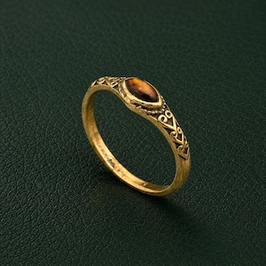 Op de afbeelding: Een goudkleurige metalen ring met een bruine tijgeroog-edelsteen in marquise-slijpvorm in het midden. De band heeft een ingewikkeld, verhoogd filigraanpatroon aan de zijkanten, met een kraalachtige textuur die de centrale zetting omrandt.