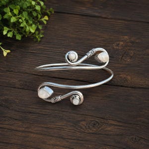May include: A silver-toned bypass cuff bracelet featuring two spiraled ends, each accented with a smooth, round, milky white gemstone. The metal band has a polished finish with decorative wire-wrapped detailing near the stone settings.