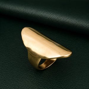 May include: A gold-toned ring featuring a wide, elongated, and smooth shield-like surface that extends across the finger. The metal has a polished finish with a slight curve, creating a bold, solid, and continuous form that tapers toward the band.