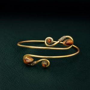 May include: A gold-toned metal cuff bracelet featuring a bypass design with two spiraling ends. Each end is accented with a small, round, brown gemstone and a larger, teardrop-shaped, brown gemstone, both displaying a chatoyant, tiger-eye effect.