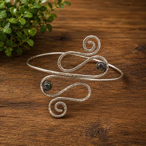 Peut inclure: Un bracelet manchette en métal argenté avec une surface texturée et deux motifs en fil métallique en forme de spirale ouverte. Deux petites pierres rondes aux reflets irisés sombres sont serties dans les courbes du fil métallique.