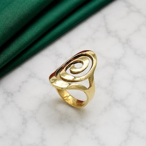May include: A gold-toned ring featuring an openwork design with a prominent central spiral motif. The band is smooth and circular, connecting to the larger, rounded top section that showcases a continuous, swirling metal pattern throughout.