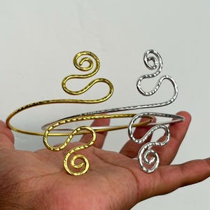 Può includere: Due bracciali da braccio, uno color oro e uno color argento, caratterizzati da una texture martellata e un design a filo continuo che forma due spirali simmetriche alle estremità. Le fasce metalliche sono curvate per adattarsi al braccio.
