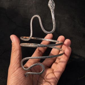 Può includere: Un bracciale da braccio in metallo color argento a forma di serpente avvolto. Il design presenta un motivo testurizzato simile a squame che copre l'intera lunghezza del corpo del serpente, con una testa dettagliata in alto e una coda curva.
