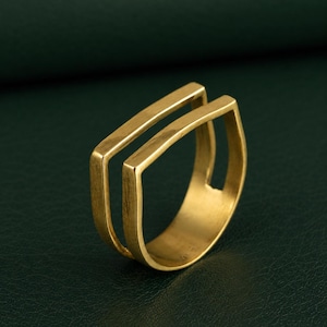 Peut inclure: Une bague dorée présentant un design à double anneau avec une finition lisse et polie. La bague se compose de deux barres métalliques rectangulaires parallèles qui se rejoignent à la base, créant une structure géométrique ouverte et uniforme.