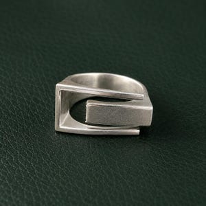 Könnte beinhalten: Ein silberfarbener Ring mit einem geometrischen, offenen Design und einem rechteckigen Balkenelement. Das Metall hat eine matte, gebürstete Oberfläche und die Struktur besteht aus zwei parallelen Bändern, die durch ein Segment verbunden sind.