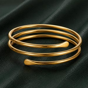 Op de afbeelding: Een goudkleurige wikkelarmband van metaal met een doorlopend spiraalontwerp met drie cirkelvormige lussen. De armband heeft een gladde, gepolijste afwerking en eindigt aan beide kanten in licht afgeplatte, afgeronde uiteinden.