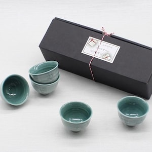 May include: Four small, round, light teal ceramic cups with a glossy finish arranged near a rectangular black gift box. The box is tied with thin red and white twine and features a white label with small gift box illustrations and Japanese text.