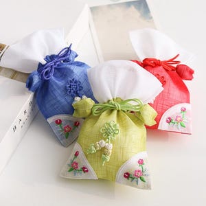 May include: Three small fabric drawstring pouches in blue, lime green, and red. Each pouch features a white top section, a textured body, and white triangular corner accents embroidered with small pink and green floral patterns.
