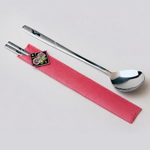 May include: A set of stainless steel chopsticks and a long-handled spoon resting on a rectangular pink fabric sleeve. The sleeve features a small, square black patch with a colorful embroidered butterfly design at the top edge.