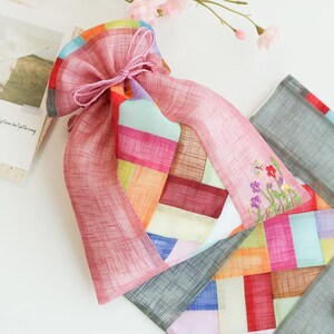 Op de afbeelding: Een buidel met trekkoord van doorschijnende, gestructureerde stof met een patchworkontwerp van veelkleurige rechthoekige panelen in roze, oranje, geel, blauw en grijs. De buidel is dichtgetrokken met roze koorden en versierd met bloemenborduurwerk.