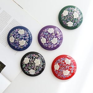 May include: Five circular compact mirrors in green, blue, purple, black, and red. Each features a central inlay of white floral motifs surrounded by iridescent, shimmering mother-of-pearl leaves and stems set against a glossy, solid-colored base.