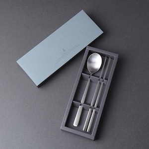 May include: A stainless steel spoon and a pair of chopsticks set inside a dark gray rectangular tray. The set includes a matching light blue lid with a small tree icon and printed text, designed for organized storage of the metal utensils.