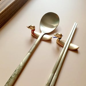 May include: A gold-toned metal spoon and a pair of chopsticks resting on two gold-toned duck-shaped holders. The spoon handle is engraved with the word KOREA and a floral pattern. One duck holder has a red neck accent, and the other has a blue one.