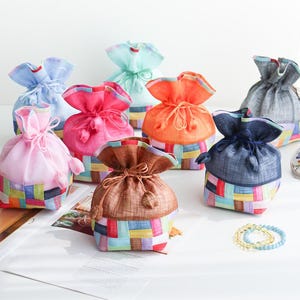Puede incluir: Una colección de ocho bolsas con cordón que presentan una sección superior de color sólido y una base construida a partir de un mosaico de cuadrados de tela multicolores. Cada bolsa se cierra con lazos de tela a juego y pequeños borlas.